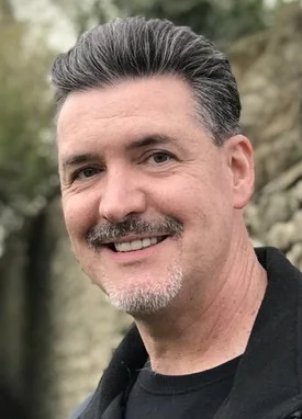 Professional headshot of smiling man in black shirt against blurred outdoor background.